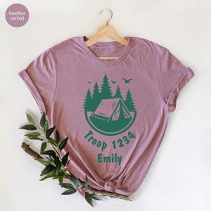 Może przedstawiać: Koszulka w kolorze wrzosu z zielonym motywem kempingowym. Projekt zawiera namiot, drzewa i tekst "Troop 1234 Emily". Koszulka jest prezentowana na drewnianym wieszaku.