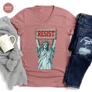 May include: A dusty rose V-neck t-shirt with a graphic of the Statue of Liberty holding a sign that reads "RESIST" in red letters. The shirt is paired with blue denim jeans and a grey sweater.