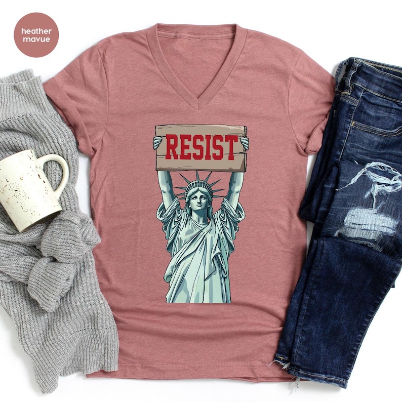 May include: A dusty rose V-neck t-shirt with a graphic of the Statue of Liberty holding a sign that reads "RESIST" in red letters. The shirt is paired with blue denim jeans and a grey sweater.