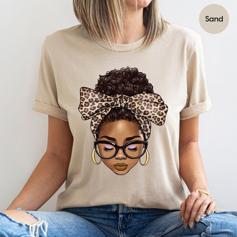May include: A beige t-shirt with a graphic of a woman with brown hair in a bun wearing a leopard print headband and glasses. The text "Sand" is in the upper right corner of the image.