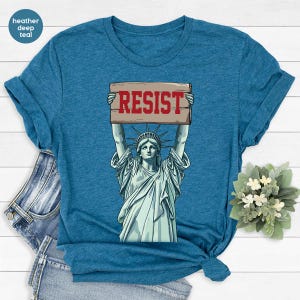 May include: Deep teal t-shirt with a graphic of the Statue of Liberty holding a sign that reads "RESIST" in red. The Statue of Liberty is in light green. The shirt is folded. The background is light. The shirt is made of a soft material.