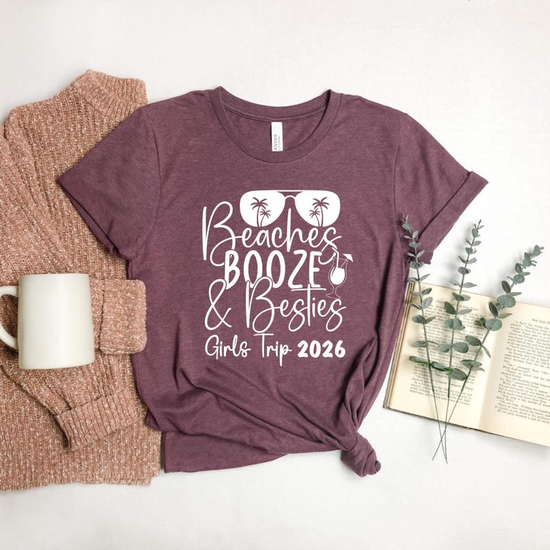 May include: A maroon t-shirt with the text "Beaches, Booze & Besties, Girls Trip 2026" in white script. The design includes sunglasses, palm trees, and a cocktail. The shirt is folded, with a mug and sweater visible.