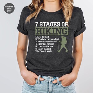 May include: A heather dark grey t-shirt featuring the white and olive green text 7 STAGES OF HIKING followed by a numbered list of seven phrases and a graphic silhouette of a hiker with a backpack and trekking pole on the right side.