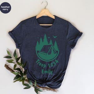 Może przedstawiać: Granatowa koszulka z zieloną grafiką kempingową. Projekt zawiera namiot, drzewa i ptaki w okręgu, z napisem "Troop 1234 Emily". Koszulka jest zawieszona na wieszaku.