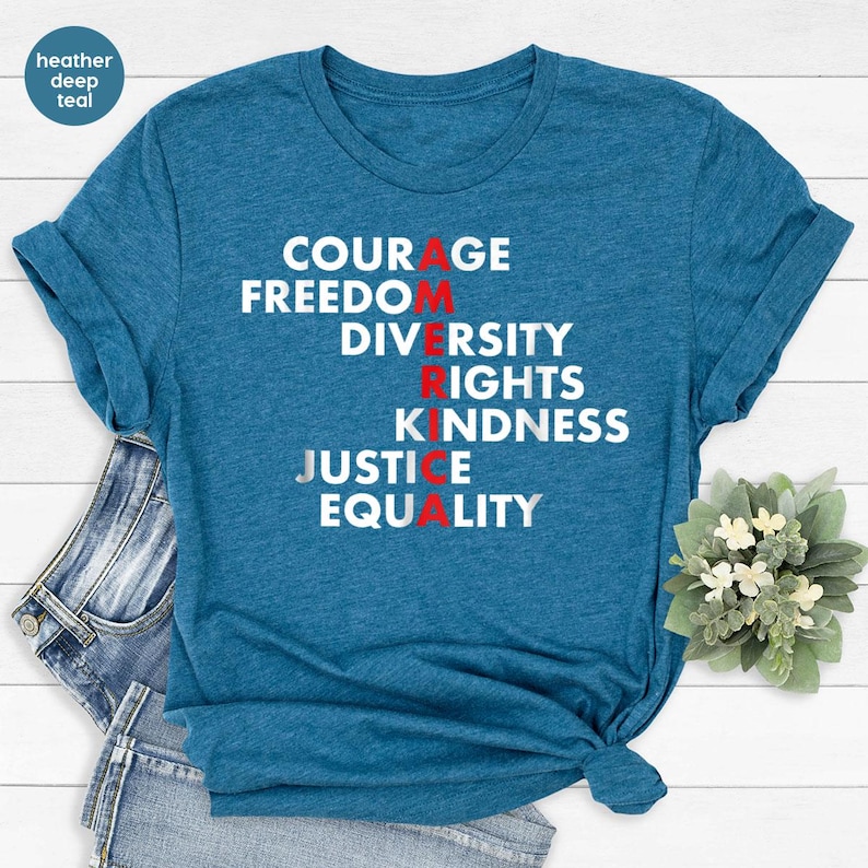 May include: A teal t-shirt with the words COURAGE, FREEDOM, DIVERSITY, RIGHTS, KINDNESS, JUSTICE, and EQUALITY printed in white. The letters 'A', 'R', and 'E' are in red. The shirt is folded and placed on a pair of blue jeans.