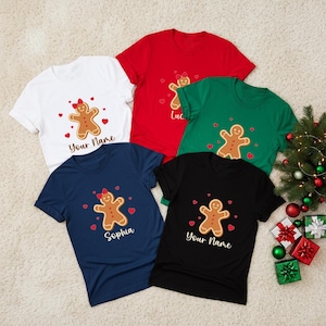 May include: Five short-sleeved t-shirts in white, red, green, navy blue, and black. Each shirt features a central gingerbread person graphic with a red bow, surrounded by small red hearts and the printed text Your Name or Sophia.