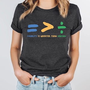 May include: A dark grey t-shirt with a graphic of an equals sign, a greater than sign, and a division sign. The text "EQUALITY IS GREATER THAN DIVISION" is printed below the graphic.