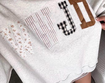 May include: A light grey heathered sweatshirt featuring the letters WWJD as large fabric appliqués. Each letter has a distinct pattern: the first W is white with small floral prints, the second W has brown stripes, the J is black and white gingham, and the D is solid brown.