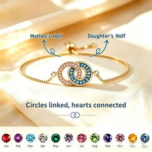 Puede incluir: Pulsera dorada con dos círculos entrelazados, uno rosa y otro azul, adornados con piedras preciosas. El texto dice "Mother's Half" y "Daughter's Half". La pulsera tiene una cadena fina y un cierre deslizante. También se ve el texto "Circles linked, hearts connected".
