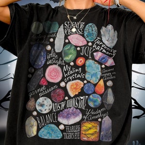 Op de afbeelding: Zwart T-shirt met een grafische print van verschillende kristallen en hun bijbehorende betekenissen, waaronder seleniet reinigen, ioliet inzicht, turquoise harmonie, granaat passie, amethist wijsheid en tijgeroog onverschrokken.