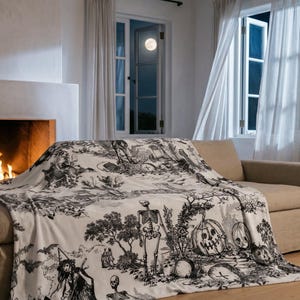 May include: A cream-colored throw blanket with a black Halloween-themed toile pattern is draped over a beige sofa. The design includes skeletons, witches, pumpkins, and bats. A fireplace with a burning fire is in the background.