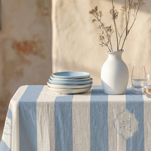 Puede incluir: Una mesa puesta con un mantel a rayas azules y blancas, una pila de platos azul claro, un jarrón blanco con flores secas y dos vasos de agua. El mantel tiene un emblema decorativo.