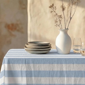 Peut inclure: Une table dressée avec une nappe à rayures bleues et blanches. Une pile d'assiettes grises, un vase blanc avec des fleurs séchées et deux verres d'eau sont posés sur la table. La scène est baignée de soleil.
