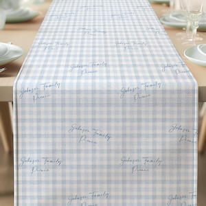 May include: A light blue and white checkered table runner with the text "Johnson Family Picnic" in a cursive font. The runner is placed on a wooden table set for a meal, with white plates and silverware.
