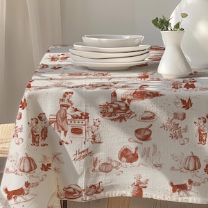 Puede incluir: Una mesa decorada para Acción de Gracias, con un mantel blanco adornado con un patrón rojo y blanco de escenas de cocina, calabazas y pavos. Platos y cuencos de cerámica blanca apilados sobre la mesa, junto a dos jarrones blancos con flores. Una silla de madera a la izquierda.