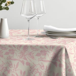 May include: A dining table set with a floral patterned tablecloth in cream and pink. Two clear wine glasses, white plates, and a folded napkin are on the table. A vase with flowers and a decorative pinecone complete the setting.