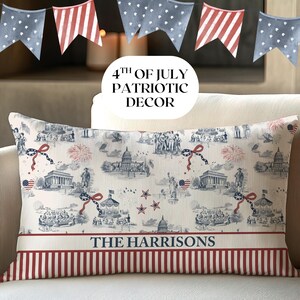 May include: Rectangular decorative pillow with a patriotic theme. The pillow features a cream-colored background with blue and red illustrations of American landmarks and symbols. The bottom has a red and white striped border, with the text "THE HARRISONS".