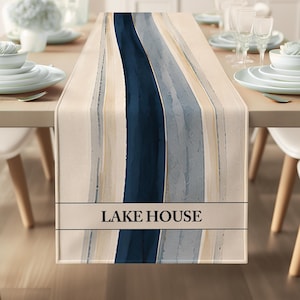 Puede incluir: Un camino de mesa rectangular con un diseño abstracto vertical de bandas en azul marino, gris claro y beige. La sección inferior incluye una franja horizontal con el texto LAKE HOUSE impreso en una fuente oscura y negrita.