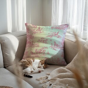 May include: A cushion with a pink and white toile pattern depicting horses and figures on a gradient background of pink, yellow, and blue. A cat is sleeping on a beige sofa next to the cushion.