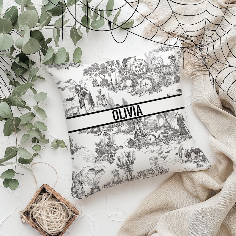 May include: A white pillow with a black and white Halloween-themed print. The print features skeletons, pumpkins, bats, and other spooky imagery. The pillow has a black band across the bottom with the name "Olivia" printed in white.