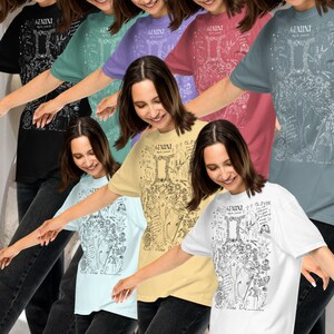 Op de afbeelding: Een groep mensen draagt T-shirts met een grafisch ontwerp van het sterrenbeeld Tweelingen. De shirts zijn in verschillende kleuren, waaronder zwart, blauw, paars, roze, geel en wit. Het grafische ontwerp toont een gedetailleerde illustratie van het sterrenbeeld Tweelingen met tekst die de eigenschappen van het sterrenbeeld Tweelingen beschrijft.