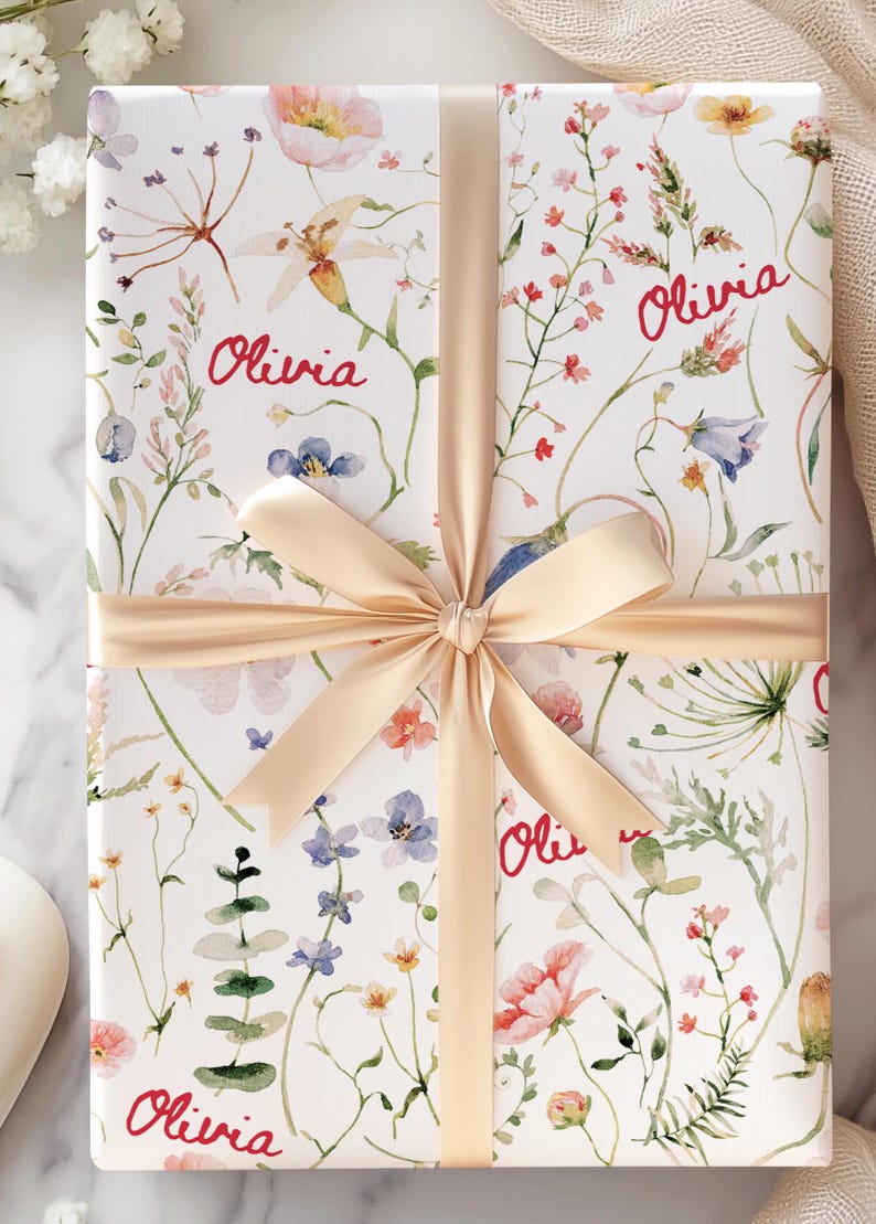 May include: A white gift box with a floral pattern and the name "Olivia" repeated four times. The box is tied with a beige satin ribbon.