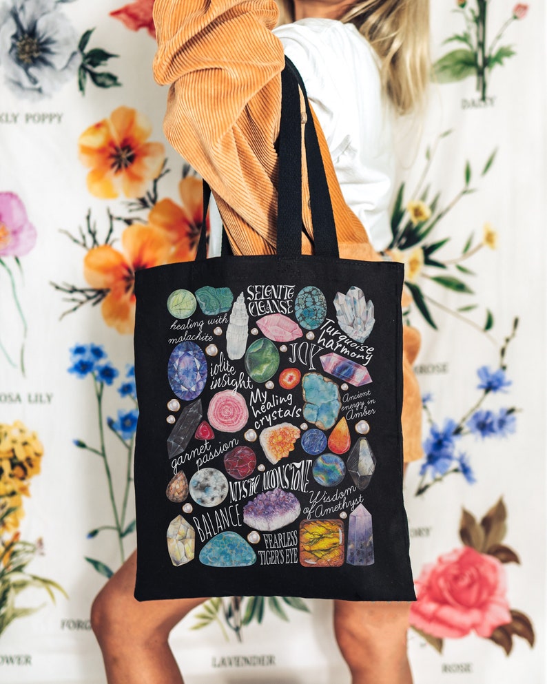 Puede incluir: Una bolsa de lona negra con un dise&ntilde;o colorido que presenta varios cristales y sus significados asociados. Los cristales incluyen selenita, turquesa, amatista, ojo de tigre y m&aacute;s. El texto en la bolsa dice "Selenite Cleanse", "Turquoise Harmony", "Healing Crystals", "Wisdom of Amethyst", "Balance", "Fearless Tiger's Eye", "Iolite Insight", "Garnet Passion", "Ancient Energy in Amber", y "Joy".
