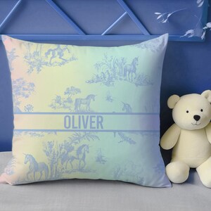 May include: Square cushion with a pastel gradient background and a blue unicorn toile pattern. The name "OLIVER" is printed in a blue band across the centre. A white teddy bear sits to the right of the cushion.