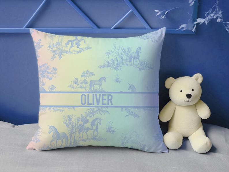 May include: Square cushion with a pastel gradient background and a blue unicorn toile pattern. The name "OLIVER" is printed in a blue band across the centre. A white teddy bear sits to the right of the cushion.