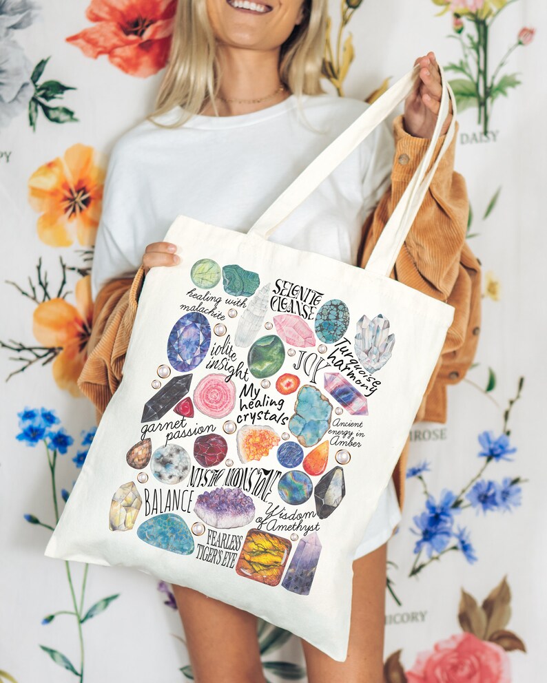 Puede incluir: Una bolsa de tela blanca con una ilustraci&oacute;n colorida de varios cristales y piedras preciosas. Los cristales est&aacute;n rodeados de texto que describe sus propiedades curativas, como "curaci&oacute;n con malaquita", "perspicacia de iolite", "mis cristales curativos", "sabidur&iacute;a de amatista" y "ojo de tigre intr&eacute;pido".