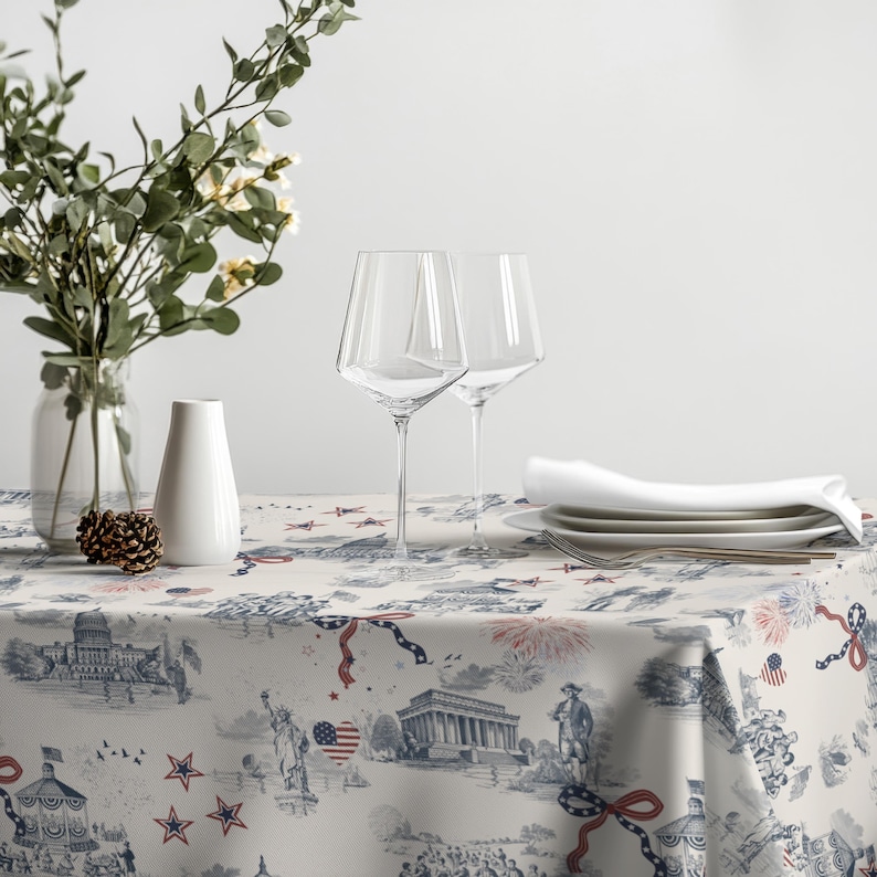 May include: A table setting featuring a white tablecloth with a blue and red patriotic design. Two clear wine glasses, white plates, and a folded napkin are on the table. A vase with flowers and a decorative pinecone are also present.