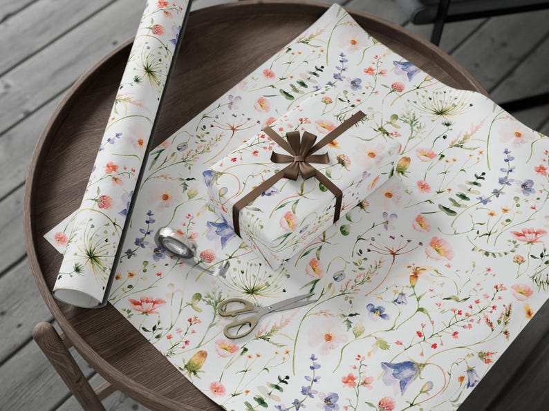 May include: White gift wrap with a watercolor floral pattern featuring pink, blue, and yellow flowers. A brown ribbon is tied around a white gift box.