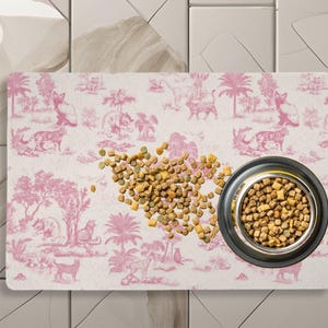 May include: A pink and white patterned pet food mat with a bowl of brown pet food. The mat has a pink and white pattern of palm trees, cats, and other jungle animals.
