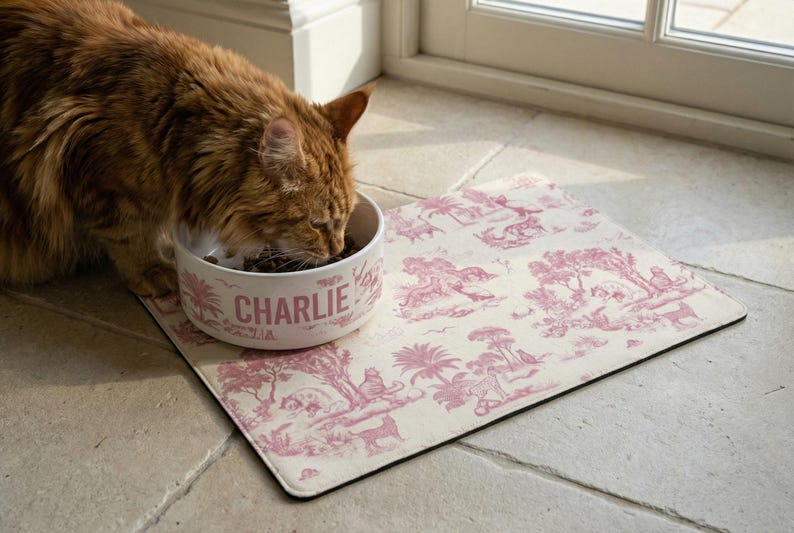 A brown cat is eating from a bowl with the name "Charlie" printed on it, placed on a pink and white patterned mat.