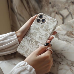 Op de afbeelding: Een witte iPhone-hoes met een gouden en wit patroon. De hoes heeft een gouden band met de tekst "DOG MOM" erop gedrukt.