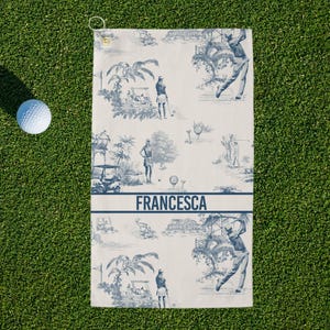 May include: Golf towel with a blue and white toile design featuring golfers and golf carts. The towel has the name "FRANCESCA" printed in blue. A white golf ball rests on the green grass next to the towel.