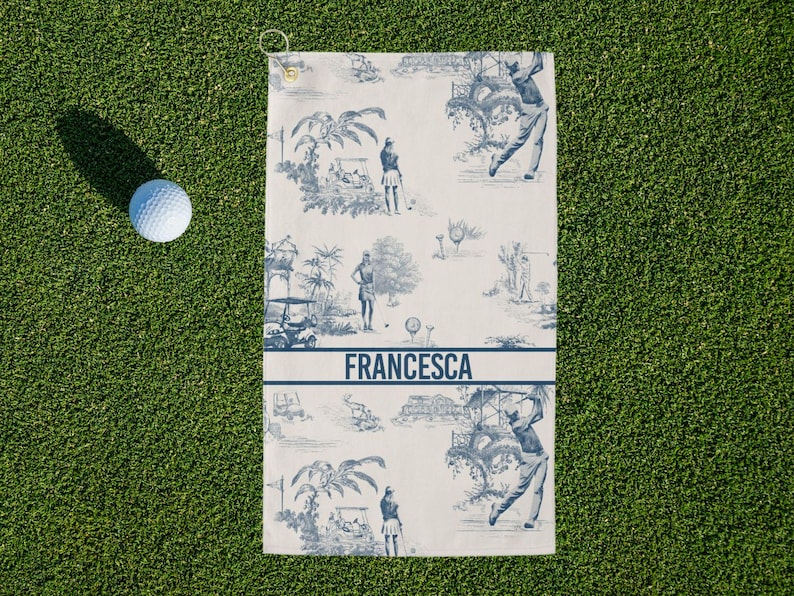May include: Golf towel with a blue and white toile design featuring golfers and golf carts. The towel has the name "FRANCESCA" printed in blue. A white golf ball rests on the green grass next to the towel.