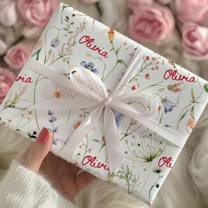 May include: White gift wrap with a floral pattern and the name "Olivia" repeated throughout. The wrap is tied with a white satin ribbon.