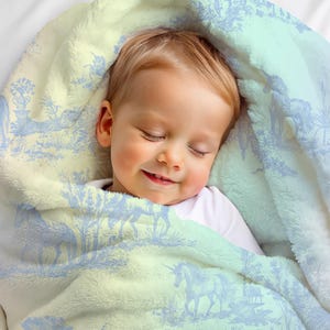 May include: A sleeping baby swaddled in a soft, plush blanket. The blanket has a light blue and cream toile pattern with unicorns and pastoral scenes. The baby is wearing a white onesie.