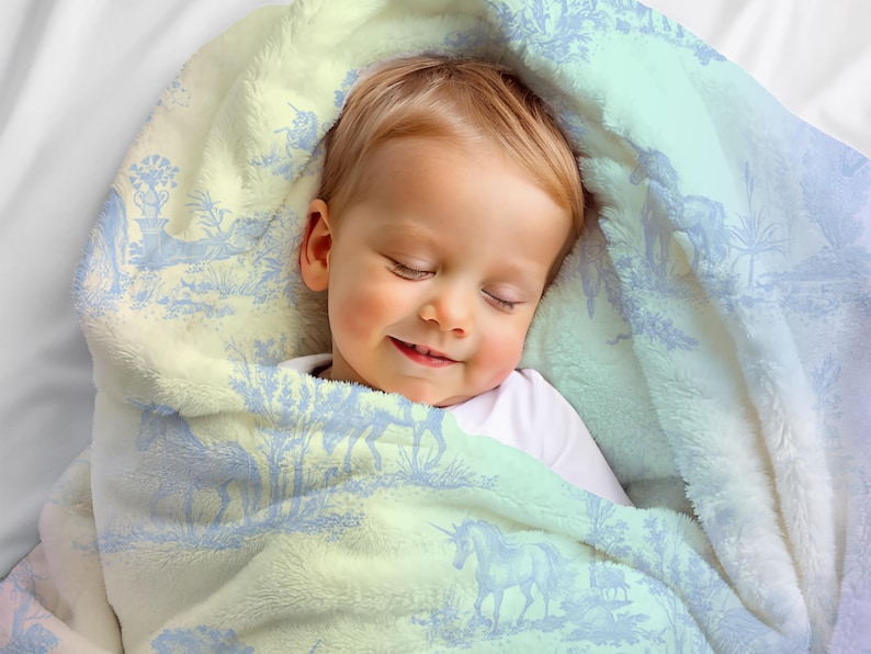 May include: A sleeping baby swaddled in a soft, plush blanket. The blanket has a light blue and cream toile pattern with unicorns and pastoral scenes. The baby is wearing a white onesie.