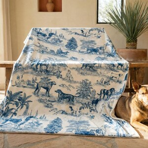 A dog is sitting on the floor next to a large blanket with a blue and white pattern of animals and nature scenes.