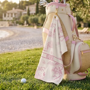 May include: A golf bag, towel, and golf ball sit on green grass. The golf bag is beige and pink with gold accents. The towel is white with a pink golf-themed design and the name "OLIVIA". A small golf bag is attached.