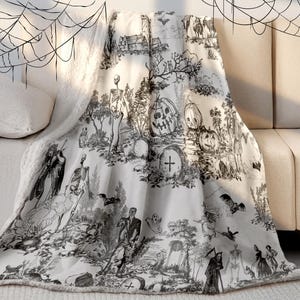 May include: A white and black Halloween-themed throw blanket draped over a sofa. The blanket features a toile design with skeletons, pumpkins, bats, and tombstones. The blanket has a soft, plush texture.