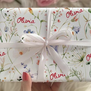 May include: White gift wrap with a floral pattern and the name "Olivia" repeated in red lettering. The gift is tied with a white satin ribbon.