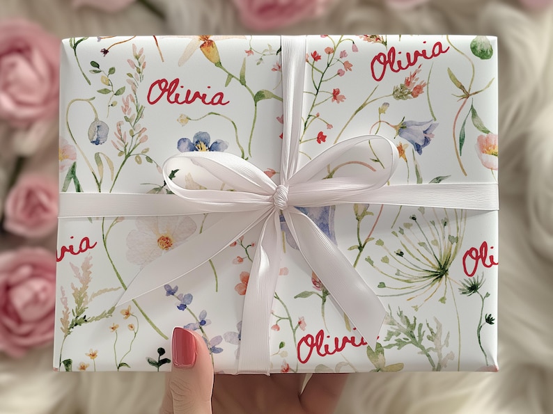 May include: White gift wrap with a floral pattern and the name "Olivia" repeated in red lettering. The gift is tied with a white satin ribbon.