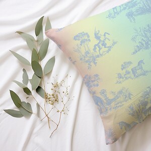 May include: A cushion with a pastel gradient and a blue toile design depicting unicorns and figures. The cushion rests on a white sheet, accompanied by eucalyptus and small white flowers.