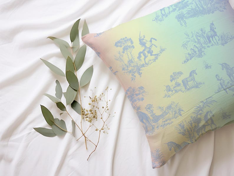 May include: A cushion with a pastel gradient and a blue toile design depicting unicorns and figures. The cushion rests on a white sheet, accompanied by eucalyptus and small white flowers.