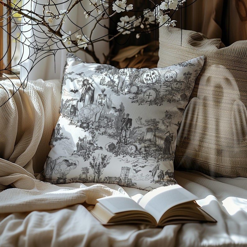 May include: A white throw pillow with a black and white print of a spooky Halloween scene. The print features skeletons, pumpkins, bats, and other spooky imagery. The pillow is sitting on a couch with a book and a throw blanket.