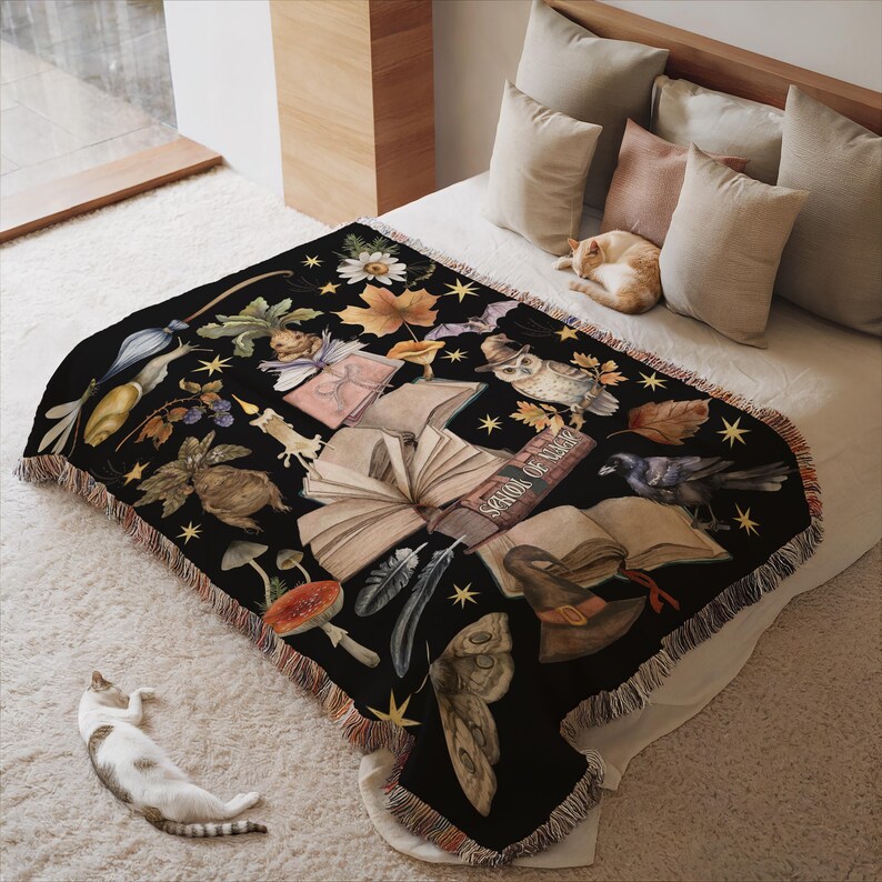 May include: A black woven throw blanket with a whimsical design featuring books, owls, mushrooms, and other autumnal elements. The text on the books reads "Spells of Magic".