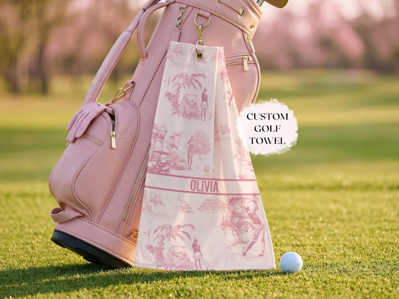 May include: A pink golf bag and a custom golf towel with a pink and white toile pattern, hanging from the bag. The towel has the name "Olivia" printed on it. A golf ball rests on the green grass.